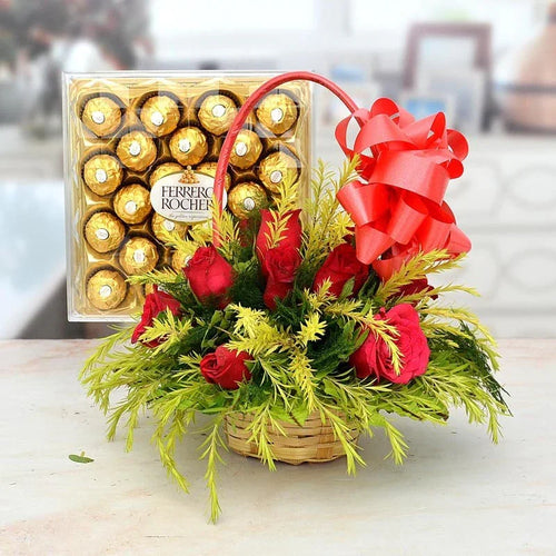 Basket of red roses with a large red bow next to a box of Ferrero Rocher chocolates on a blurred background