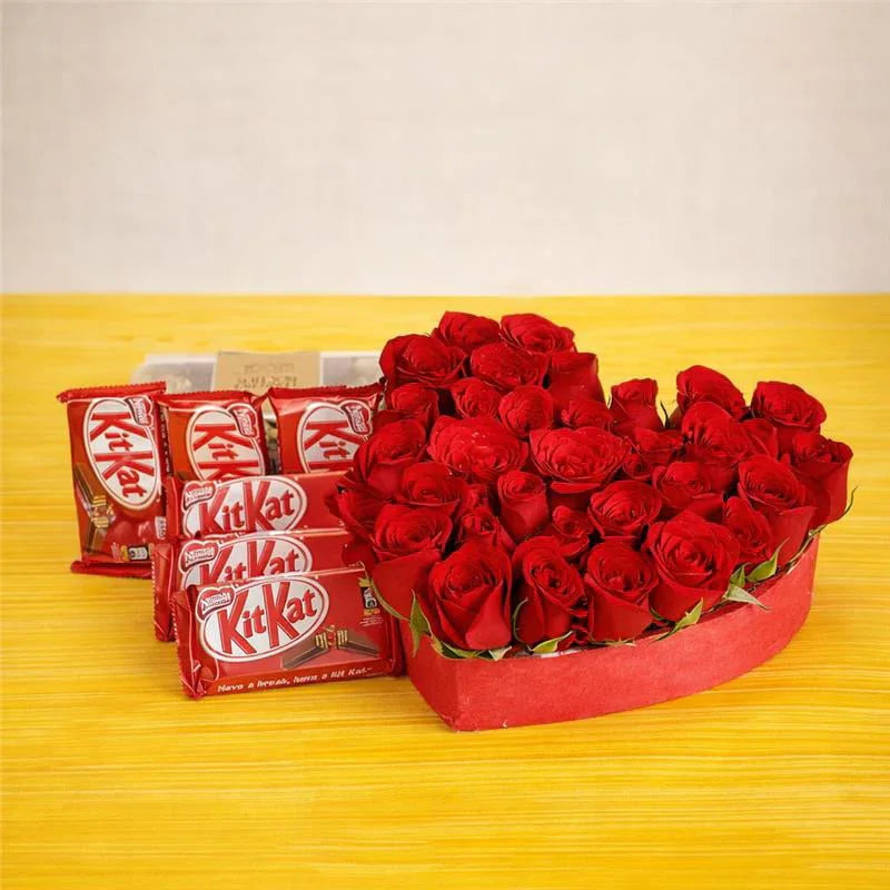 Red roses in a heart-shaped box next to KitKat chocolate bars on a yellow surface.