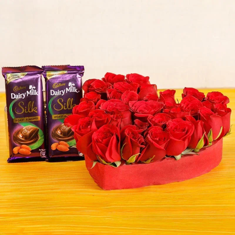 Heart-shaped arrangement of red roses with two Cadbury Dairy Milk Silk chocolate bars on a yellow surface.