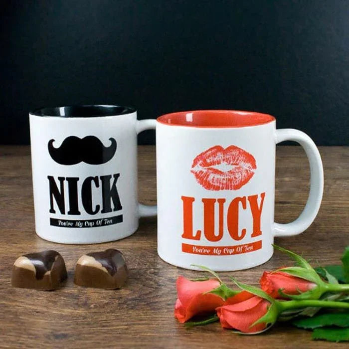 Two mugs with personalized designs and text, accompanied by candies and roses on a wooden surface.