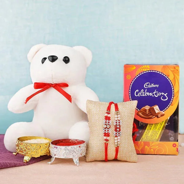White teddy bear with a red bow, two Rakhi on a pillow, and a Cadbury chocolate box on a light blue background.