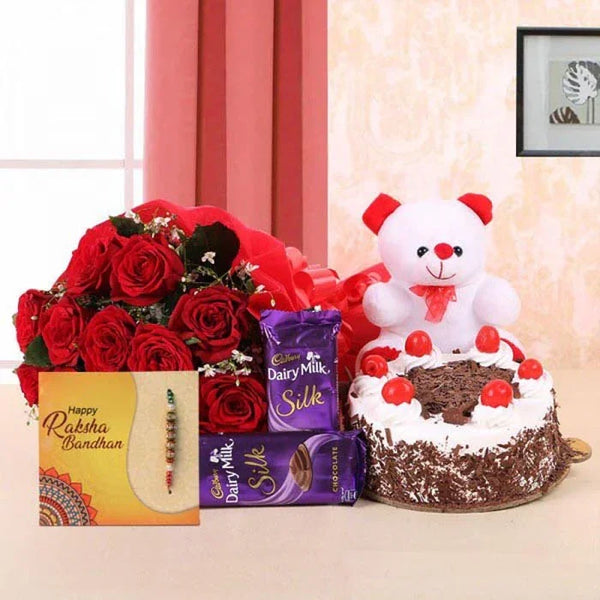 Gifting arrangement with red roses, teddy bear, cake, and Cadbury Dairy Milk Silk on a beige surface.