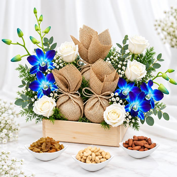 A gift arrangement in a wooden box featuring three burlap-wrapped spheres tied with twine, white roses, blue orchids, green foliage, and small white baby's breath. Three small bowls of nuts and dried fruit sit in front on a white draped background.