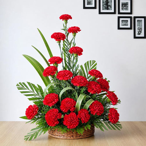Bouquet of red carnations in a woven basket on a wooden surface with a white wall background.