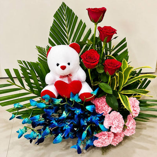 Bouquet of red roses, blue orchids, pink carnations, and a white teddy bear with a red heart on a light background.