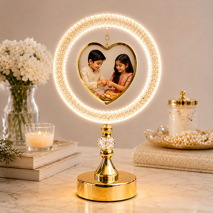 A gold LED lighted picture frame table lamp with a heart-shaped photo display and a circular ring of warm white lights, shown on a marble surface with decorative props in a styled indoor setting.