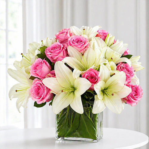 Bouquet of pink roses and white lilies in a clear vase on a white surface with a light gray curtain background.
