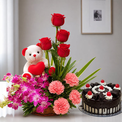 Floral arrangement with red roses, pink carnations, and a teddy bear holding a heart, next to a chocolate cake with berries on a table.