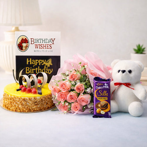 Birthday cake, bouquet of pink roses, Cadbury Dairy Milk Silk chocolate, and white teddy bear on a white surface.