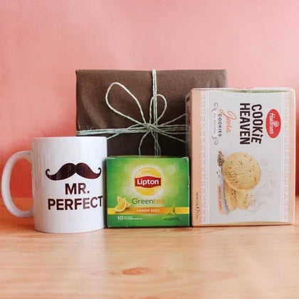 Gift set with Mr. Perfect mug, Lipton green tea, cookies, and a wrapped box on wooden table