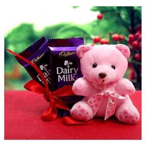 Pink teddy bear with Cadbury Dairy Milk chocolate bars on a red surface with a blurred green and red background.