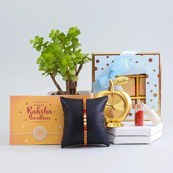 Decorative setup with a plant, gift box, clock, and black pillow with a rakhi on a light gray background.