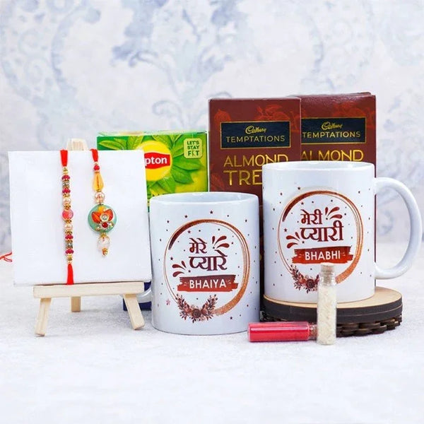 Two mugs with printed text, a box of Cadbury Temptations, a tea box, and a small decorative stand with a Rakhi on a light gray background.