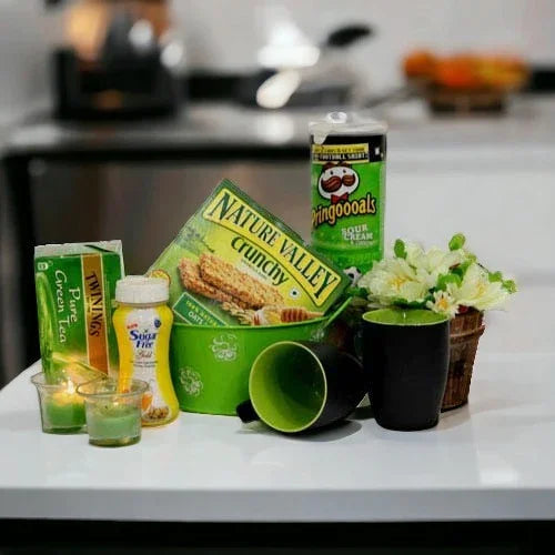 Gift hamper with snacks, green tea, mugs, flowers, and candles on a white table
