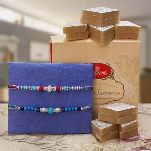 Two rakhi with a blue card and a box of Kaiju Katli savories on a table.