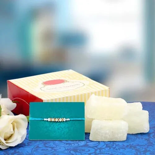 Rakhi with card, White Sweets , and flowers on a blue surface with a blurred background