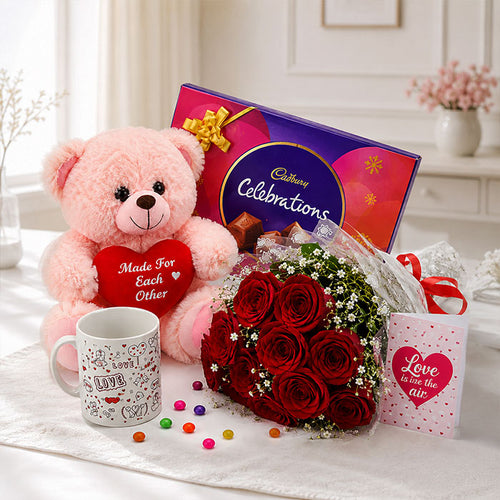 Pink teddy bear with a heart, bouquet of red roses, Cadbury chocolate box, and mug on a white surface.