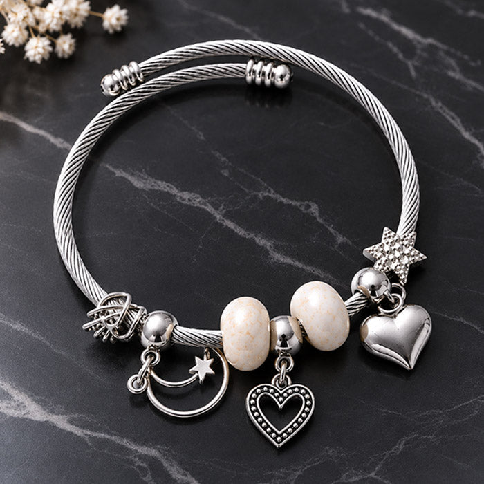 A silver-toned cable charm bracelet displayed on a dark marble surface. The bracelet features a twisted cable design, a star charm, a crescent moon with a star, a textured heart charm, a polished heart charm, and two cream-colored oval beads.