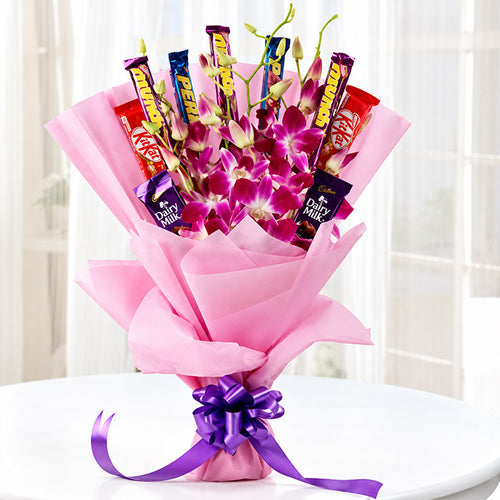 A floral bouquet gift set featuring bright pink orchids and purple blossoms arranged with assorted chocolate bars, wrapped in pink tissue paper and tied with a purple ribbon, set on a white table in front of a softly lit window.