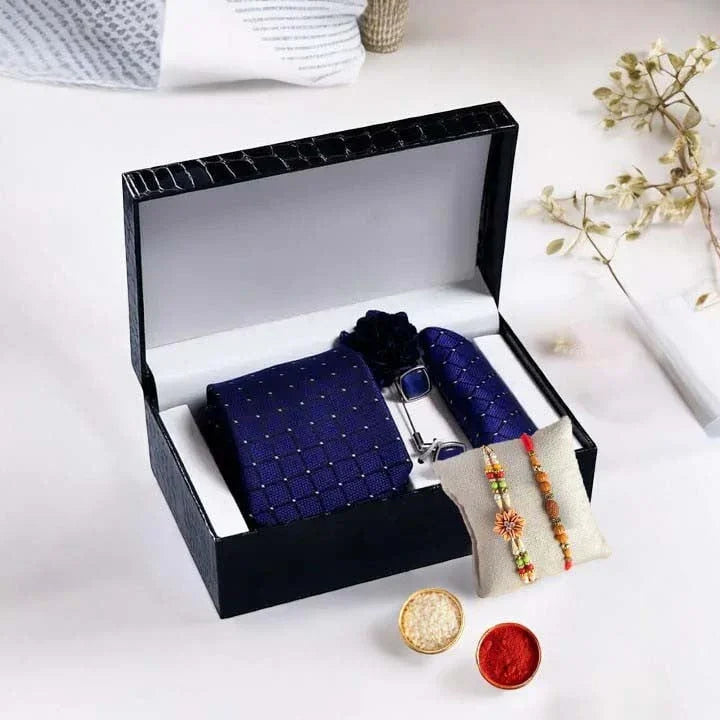 Open black gift box containing a navy blue tie, cufflinks, and a small pouch with decorative items on a white surface.