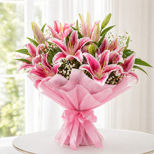 Bouquet of pink lilies wrapped in pink tissue paper on a white surface with a blurred window background.
