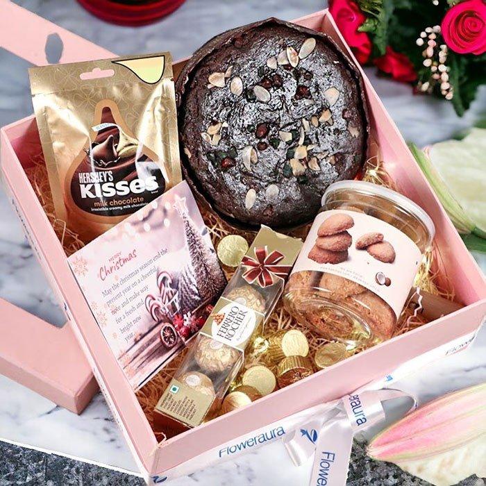Gift box with assorted treats including a chocolate cake, cookies, and a bouquet of flowers on a marble surface.