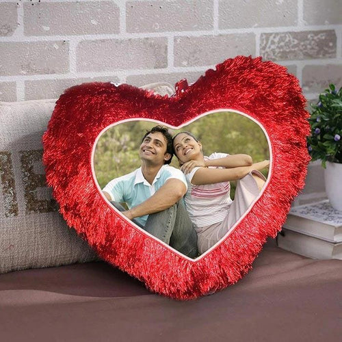 Heart-shaped red pillow with a photo of a couple embracing inside, placed on a couch against a brick wall.
