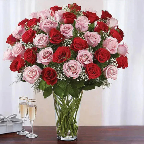 Bouquet of red and pink roses in a clear vase on a table with two champagne glasses and a gift box.