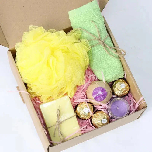 Gift box with yellow loofah, green towel, candles, and chocolates on a white background