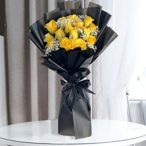 Bouquet of yellow roses wrapped in black paper with a bow on a glass table.