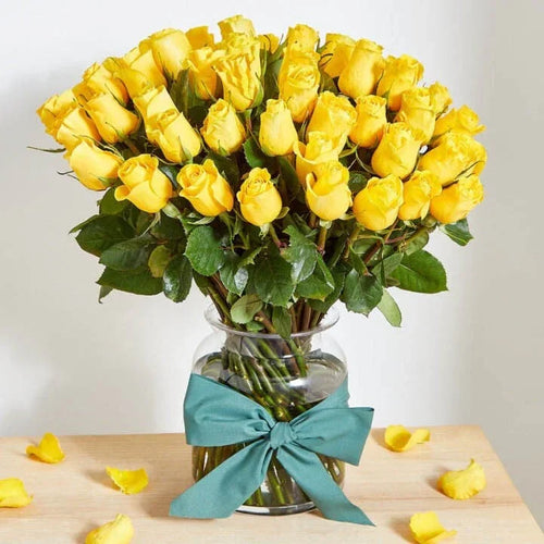 Bouquet of yellow roses in a glass vase with a teal ribbon on a wooden table.