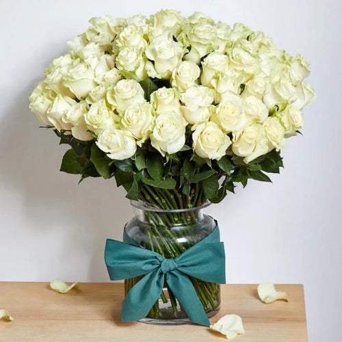 Bouquet of white roses in a clear vase with a teal ribbon on a wooden surface.