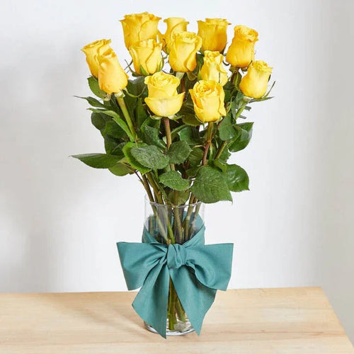 Bouquet of yellow roses in a clear vase with a teal ribbon on a wooden surface.