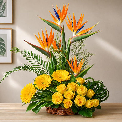 Floral arrangement with yellow flowers, green leaves, and orange and blue bird of paradise on a wooden surface.