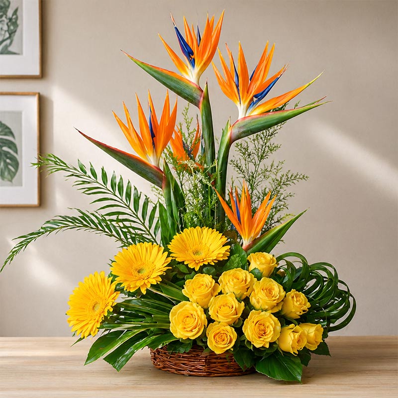 Floral arrangement with yellow flowers, green leaves, and orange and blue bird of paradise on a wooden surface.