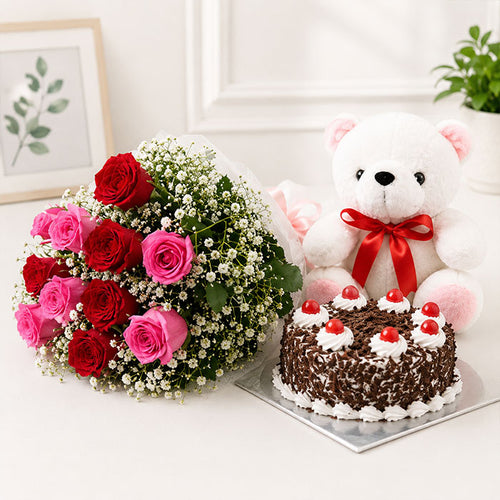 Floral arrangement with red and pink roses, a cake, and a teddy bear with a red bow on a white surface.