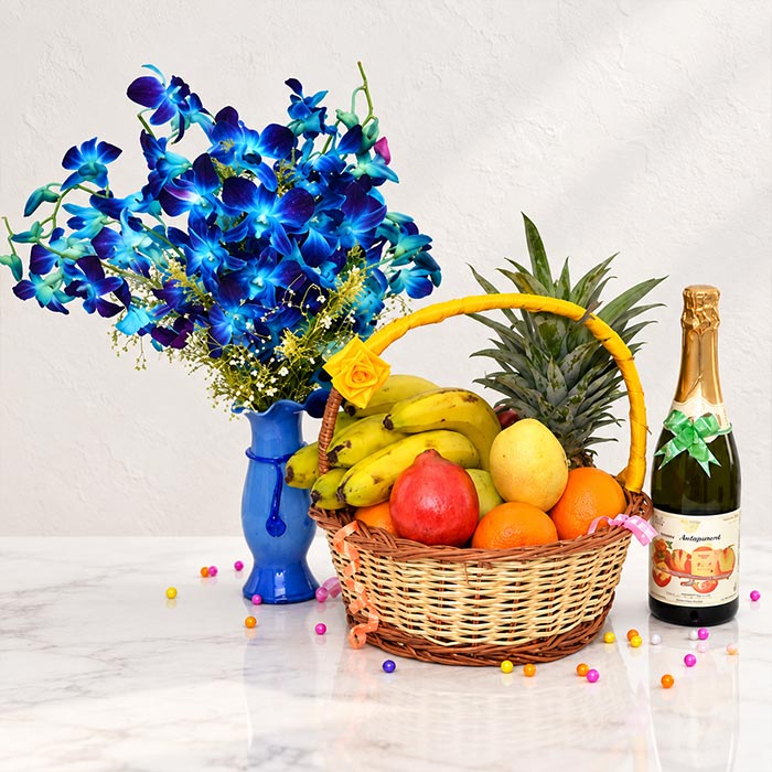 A wicker fruit basket filled with bananas, pineapple, pomegranate, oranges, and yellow fruit, arranged beside a blue vase of bright blue flowers and a champagne bottle on a white marble surface against a white wall.