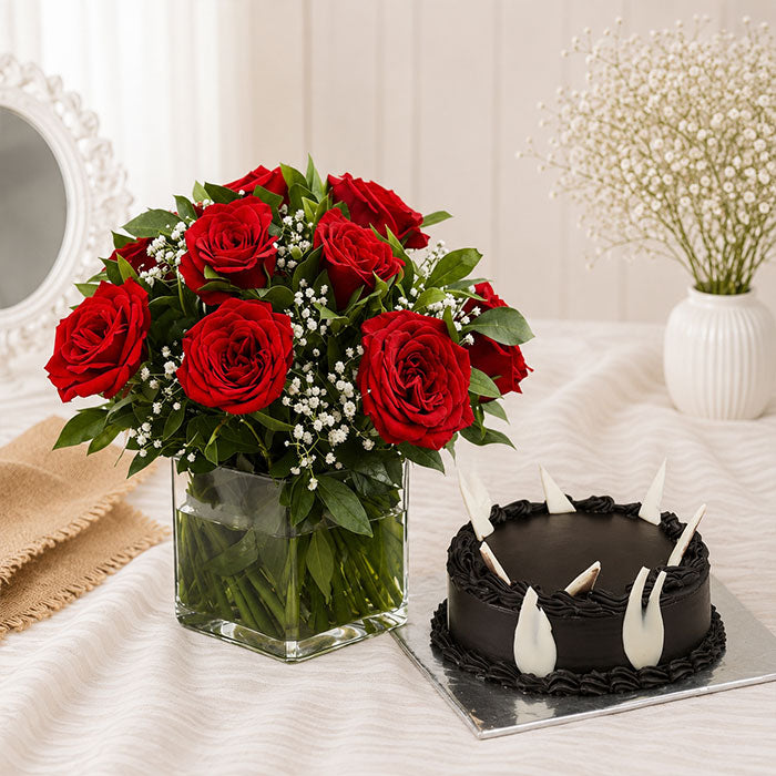 Bouquet of red roses in a clear vase next to a small chocolate cake on a white surface.
