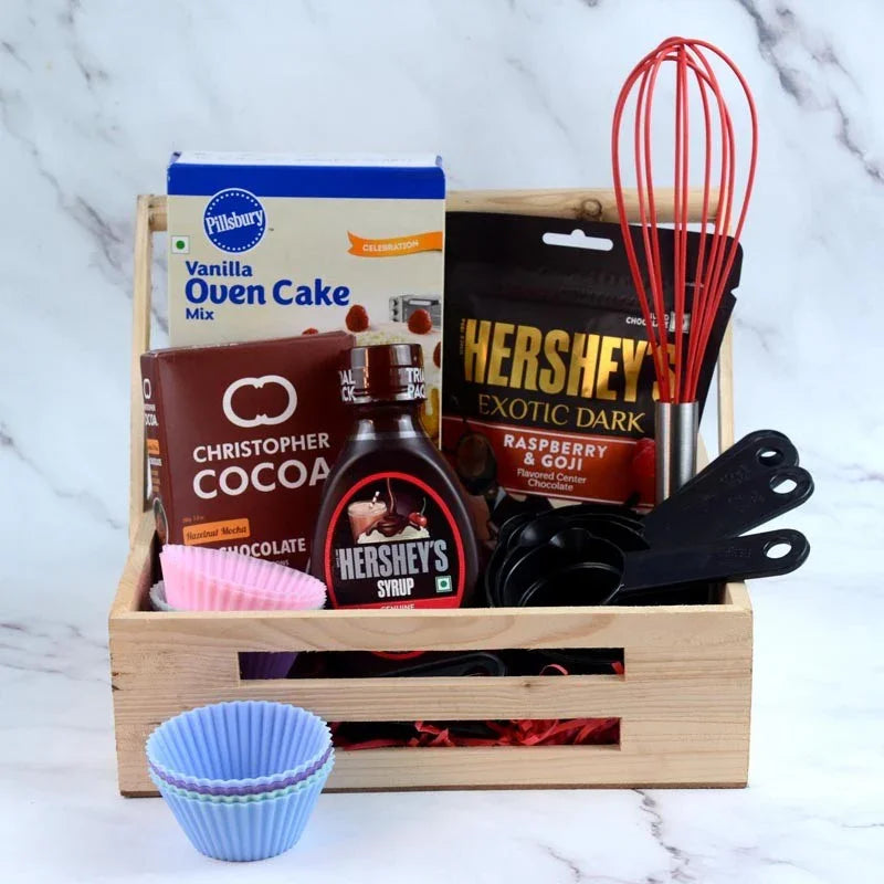 Baking kit with cake mix, chocolate, and utensils in a wooden crate on a marble surface.