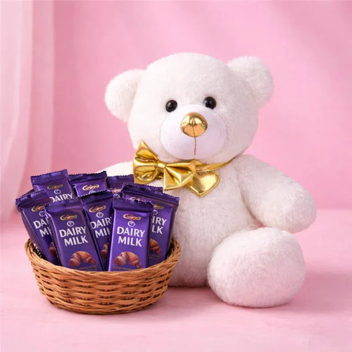 White teddy bear with a gold bow tie next to a basket of Cadbury Dairy Milk chocolate bars on a pink background