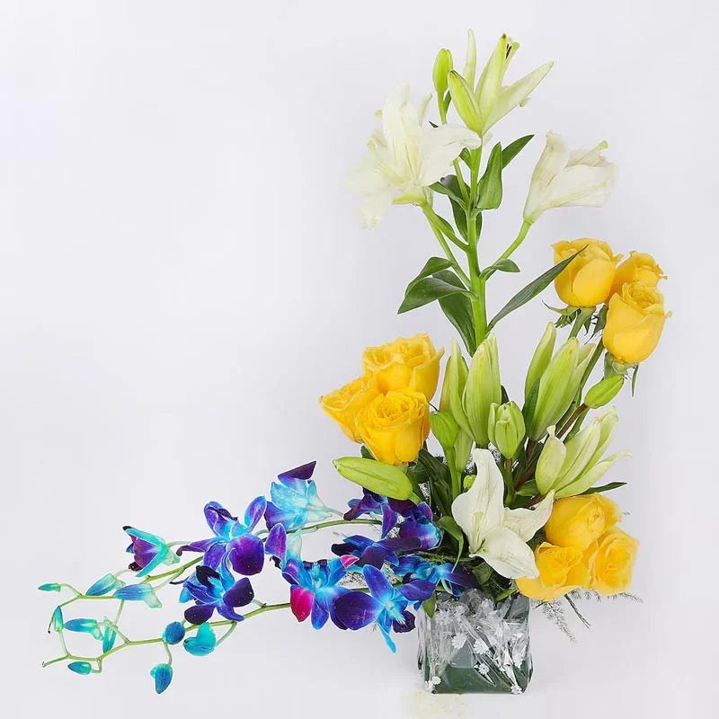 Bouquet of yellow roses, white lilies, and blue orchids in a clear glass vase on a white background