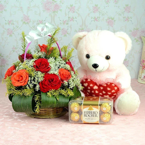 Floral arrangement with red roses, a white teddy bear, and Ferrero Rocher chocolates on a floral-patterned background.