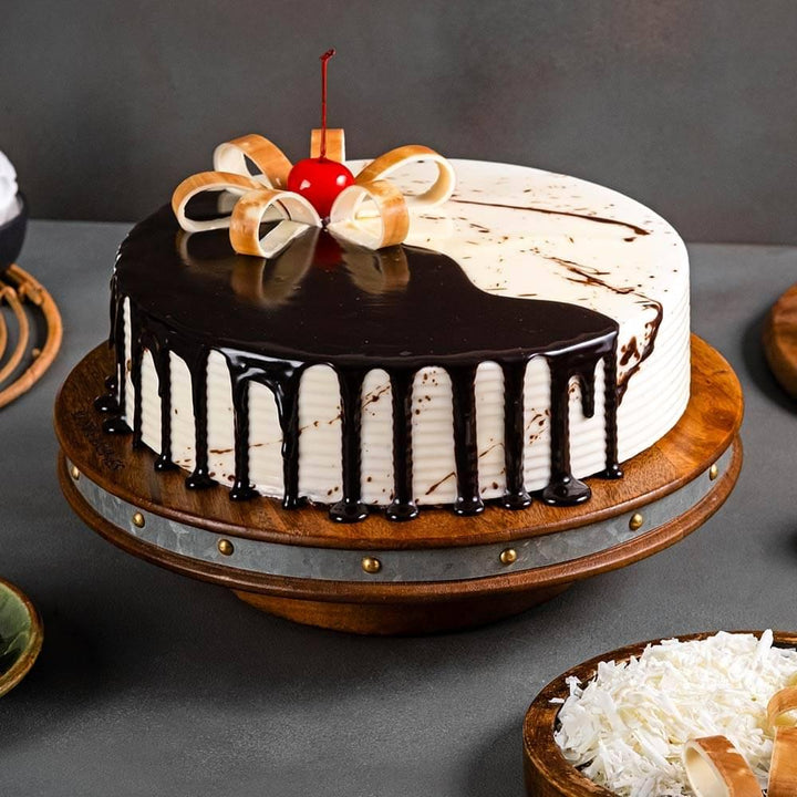 Chocolate vanilla cake with chocolate ganache drip, cherry, and white chocolate garnish on a wooden stand
