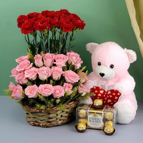 Basket of red and pink roses with a pink teddy bear holding a heart-shaped chocolate box on a green background.