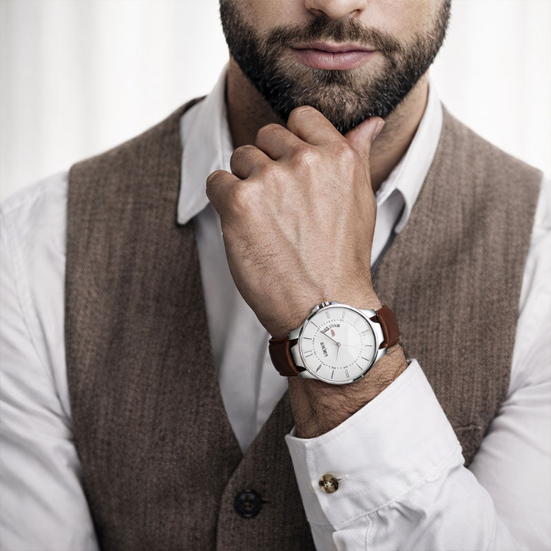 Man wearing a watch with a brown leather strap and white dial, dressed in a white shirt and brown vest.