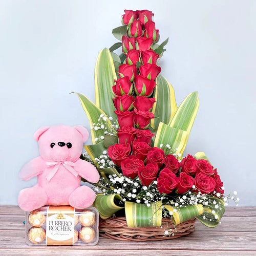 Basket of red roses, pink teddy bear, and chocolates on a wooden surface with a light gray background