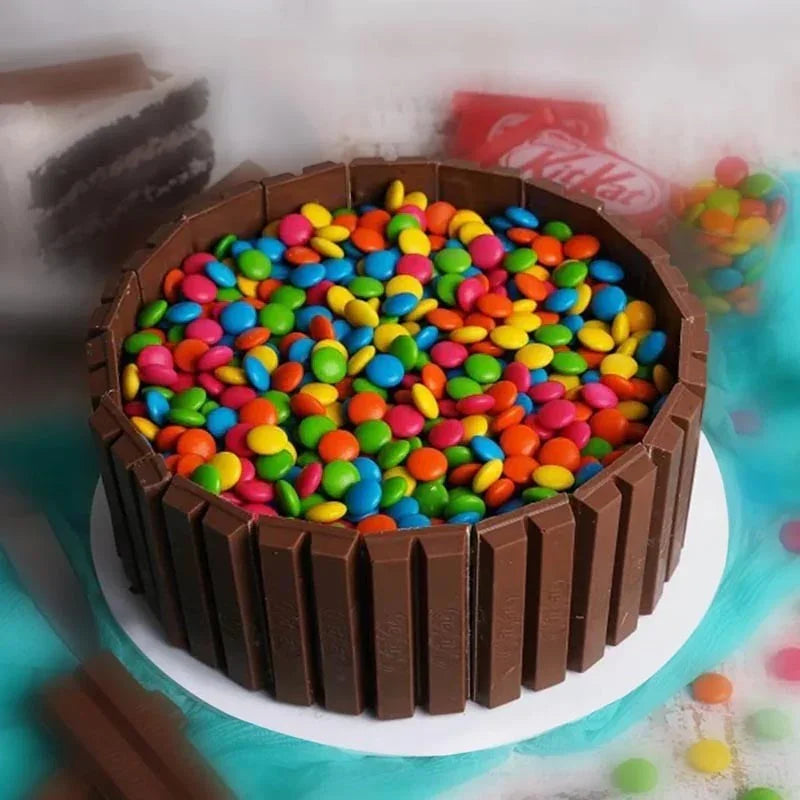Chocolate cake with colorful candy pieces on a white plate, surrounded by KitKat bars and candy.