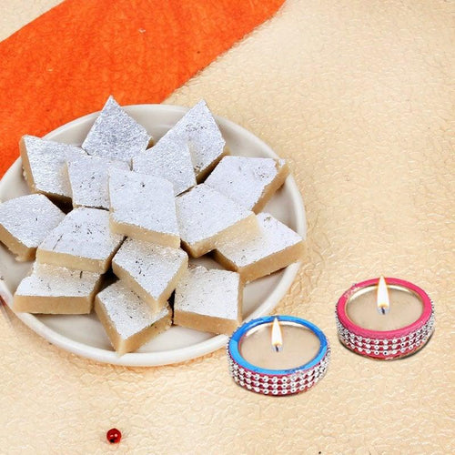 Plate of square-shaped dessert with powdered sugar on a beige surface, accompanied by two small Diya .