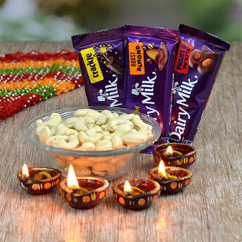 Three Dairy Milk chocolate bars with a bowl of cashews and lit diya candles on a wooden surface.