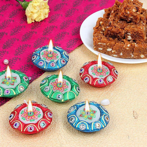 Colorful Diwali oil Diya with lit sweetson a textured surface with a plate of sweets in the background.